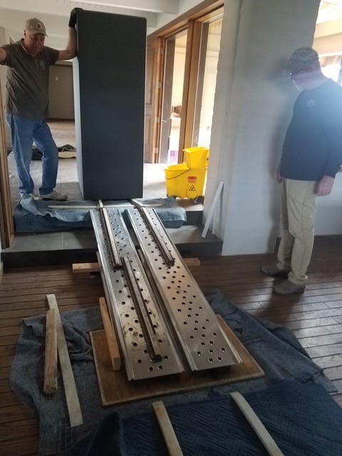 Two movers walking a dark safe over metal ramps on protected hardwood.