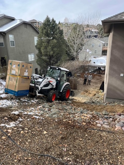 Bobcat carrying a crated safe down a steep rocky run between houses.
